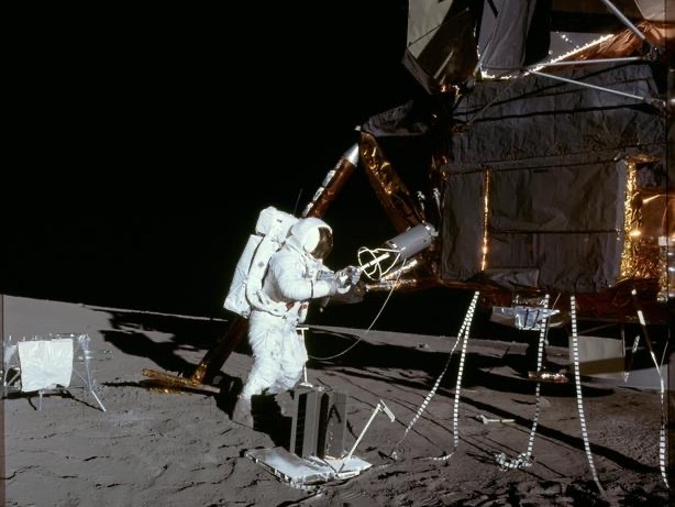 As astronaut walks on the moon's surface, holding a device